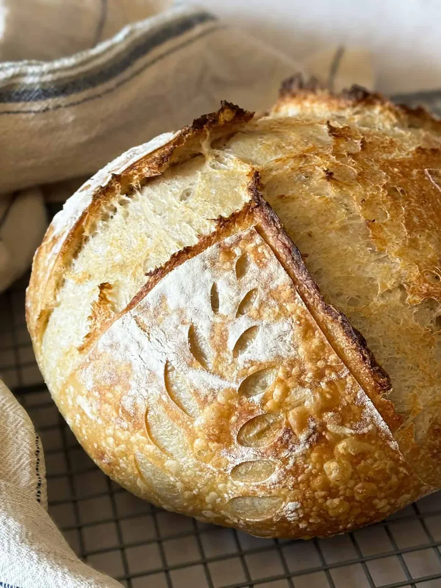A freshly baked sourdough loaf with a wheat-leaf scoring pattern, cooling on a rack beside a linen towel